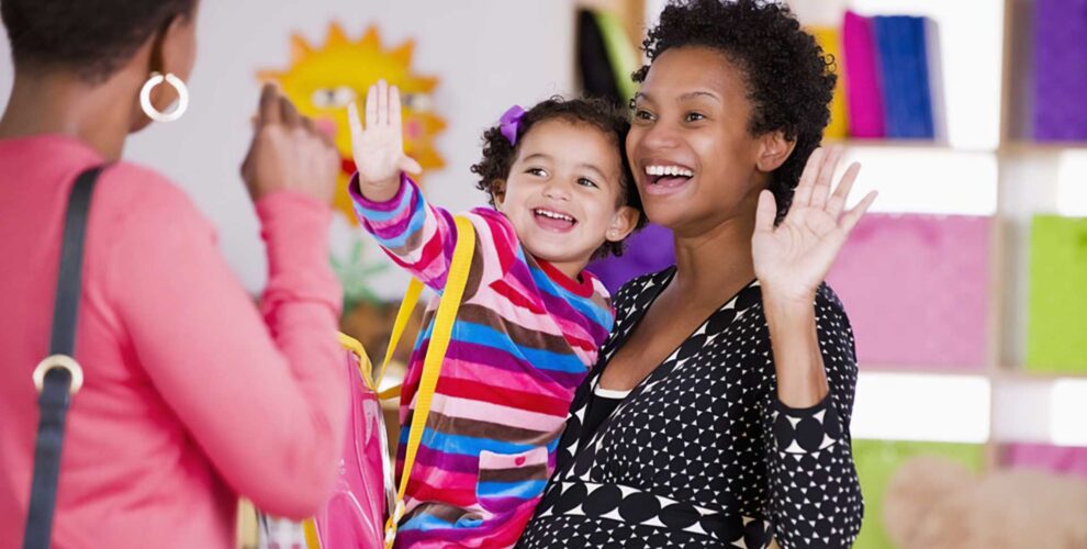 Image of teacher holding young happy child who is waving goodbye to her mother. Mother in foreground facing away from camera and waving back at child